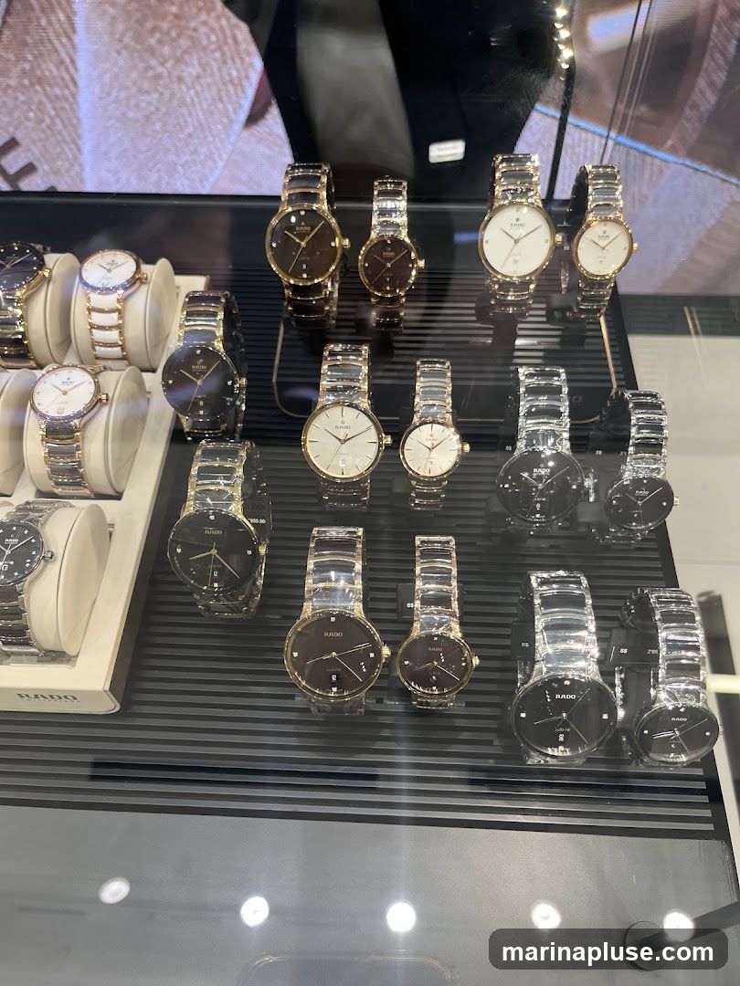 A display case filled with various Rado brand watches. The watches feature metallic bands and faces in gold, silver, and black. Some watches have diamond accents on their faces. The display is well-lit, highlighting the craftsmanship of the timepieces.