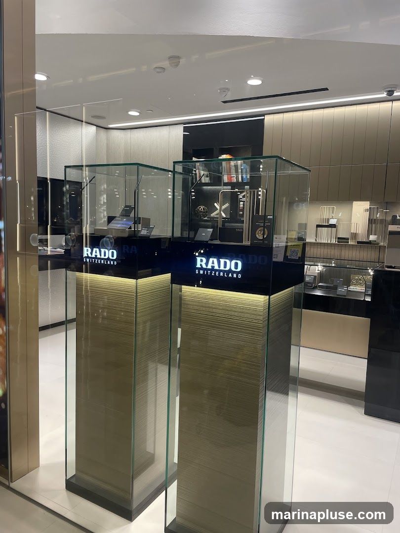 A display of Rado watches in glass cases, with the Rado logo visible on the display stands. The watches are showcased on pedestals with illuminated sides. In the background, more watches and display shelves are visible within the store.