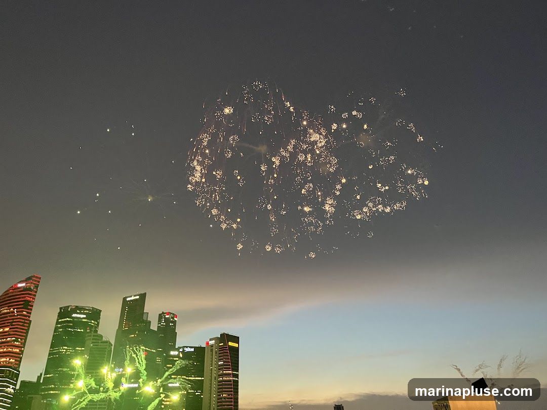 A nighttime view of a city skyline with fireworks exploding in the sky. Several tall buildings are visible, some illuminated with green lights. The sky transitions from dark gray to a lighter blue and orange near the horizon.