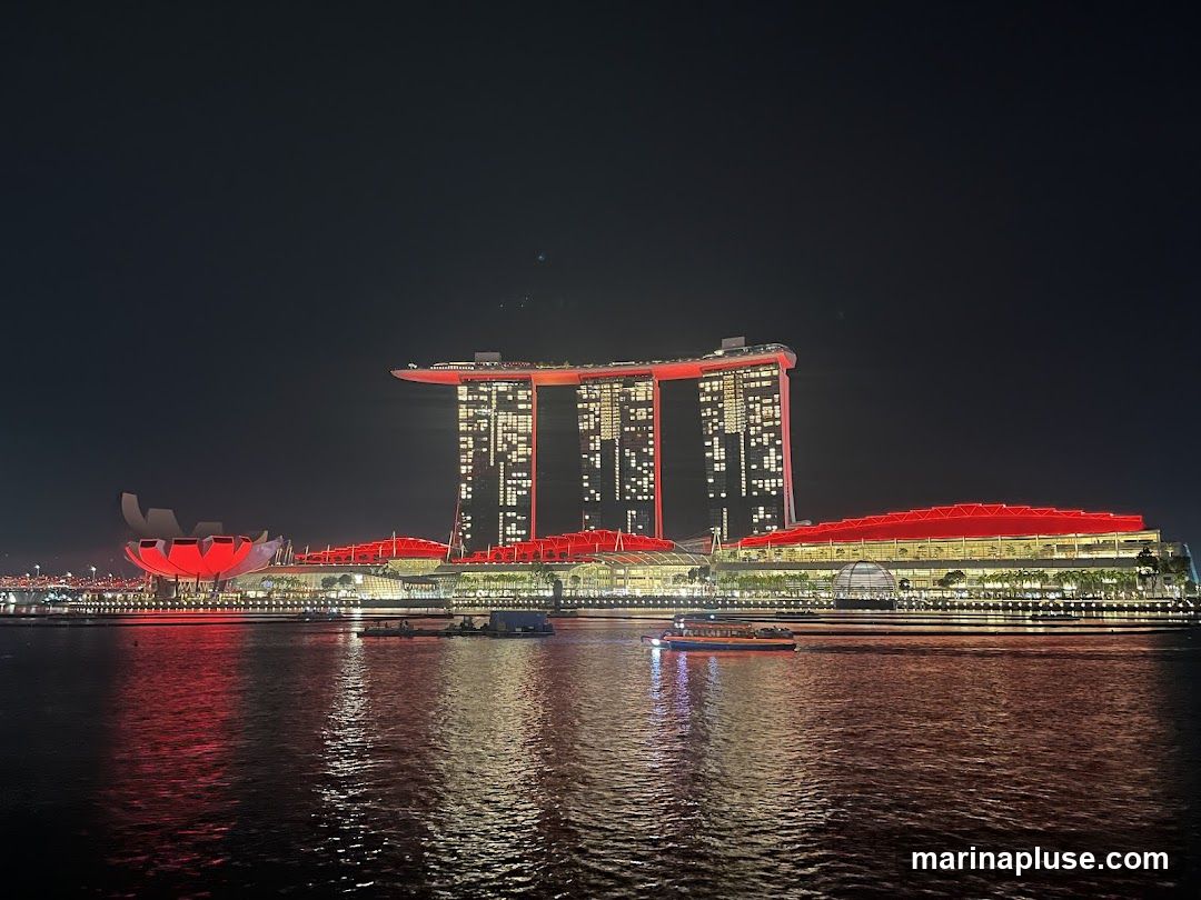 A wide shot of the Marina Bay Sands complex in Singapore at night. The iconic three towers, connected by the SkyPark, are illuminated with red lights, as is the convention center in the foreground. The lights of the buildings are reflected in the dark water of the bay. A few boats are visible on the water.