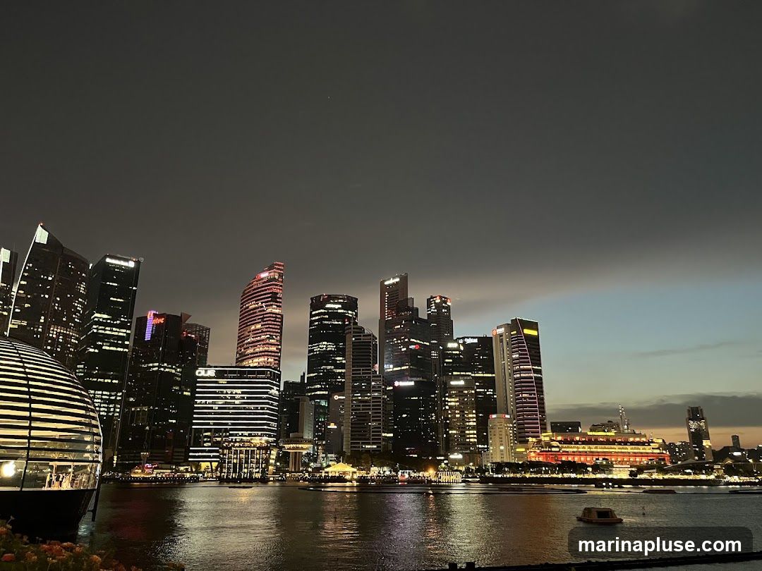 A panoramic view of a modern city skyline at dusk, with numerous illuminated skyscrapers reflecting in the calm water of a bay or river in the foreground. The sky transitions from dark gray to a lighter blue and orange hue near the horizon. A distinctive, dome-shaped building with horizontal lines is visible on the left, and a large, red-lit building complex is prominent in the mid-ground. Several boats are scattered on the water.