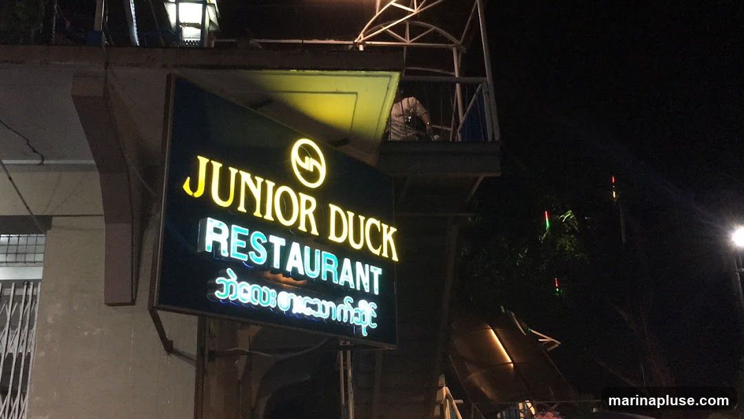 A neon sign for Junior Duck Restaurant is illuminated at night. The sign is dark blue with yellow and cyan lettering. The top line reads "JUNIOR DUCK" in yellow, with a circular logo above the 'O' in JUNIOR. The second line reads "RESTAURANT" in cyan. Below this, there is text in a different script, also in cyan neon. The sign is mounted on a dark frame and appears to be attached to the exterior of a building.