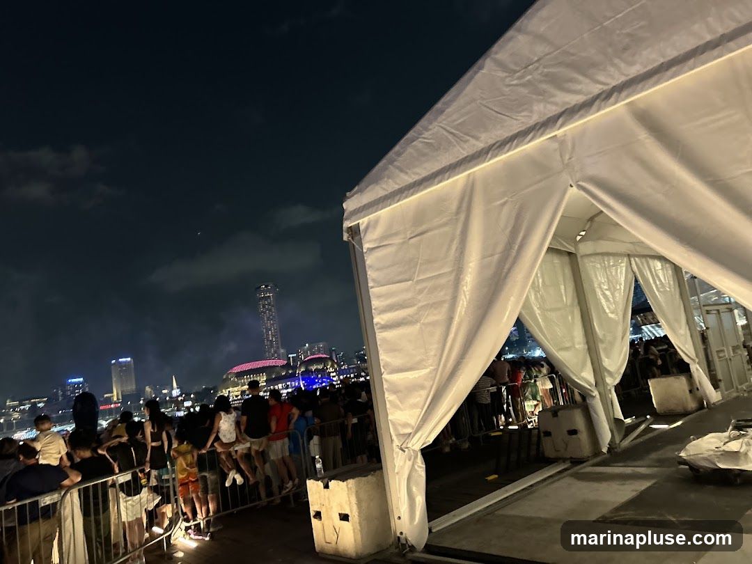 A large crowd of people are gathered outdoors at night, with a cityscape visible in the background. A large white tent structure is in the foreground, with people standing behind a barrier. The sky is dark with some clouds.