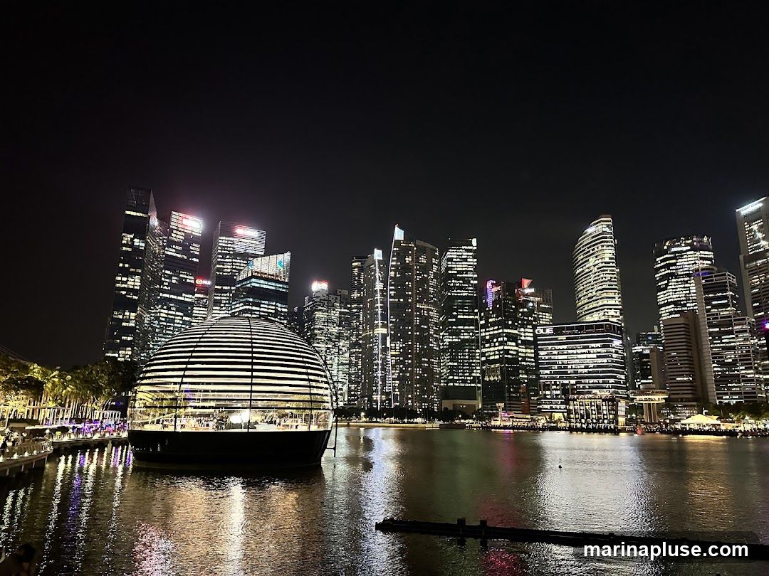 A nighttime view of a modern cityscape with a prominent spherical building on the water. The building has a glass facade with horizontal white stripes and is illuminated from within. In the background, numerous skyscrapers with lit windows form a dense urban skyline. The water in the foreground reflects the lights of the city and the spherical building. Palm trees and a walkway with people are visible to the left of the spherical structure.