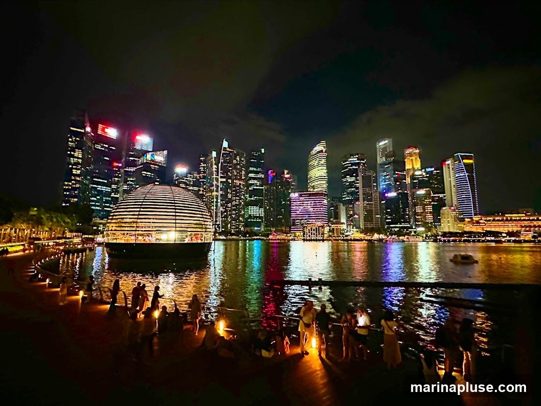 A nighttime panoramic view of Marina Bay Sands in Singapore, with the iconic Apple Store in the foreground. The domed Apple Store, illuminated from within, sits on the water, reflecting the city lights. The Singapore skyline, a cluster of modern skyscrapers, is visible in the background, all aglow with colorful lights. People are gathered along the waterfront promenade, enjoying the view. The water reflects the vibrant city lights, creating a dazzling spectacle.