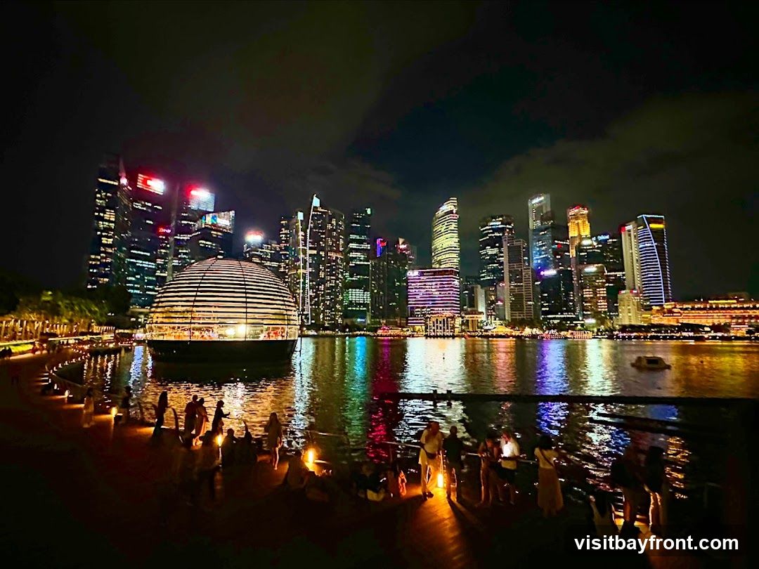 A nighttime panoramic view of Marina Bay Sands in Singapore, with the iconic Apple Store in the foreground. The domed Apple Store, illuminated from within, sits on the water, reflecting the city lights. The Singapore skyline, a cluster of modern skyscrapers, is visible in the background, all aglow with colorful lights. People are gathered along the waterfront promenade, enjoying the view. The water reflects the vibrant city lights, creating a dazzling spectacle.