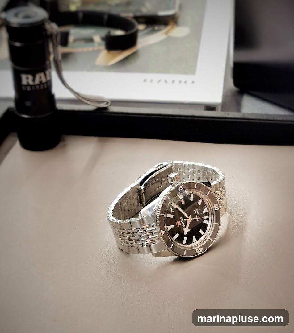 A close-up shot of a Rado watch with a black dial and silver band. To the left of the watch is a black flashlight with the Rado logo visible. In the background, a book or magazine with the Rado logo is partially visible.