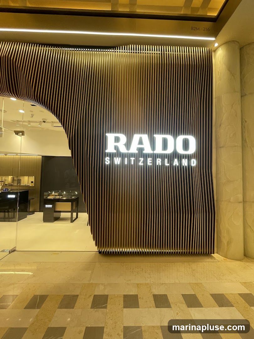 The exterior of a Rado watch store. The storefront features a modern design with vertical wooden slats and a prominent illuminated RADO SWITZERLAND logo. Through the glass entrance, display cases with watches are visible inside.