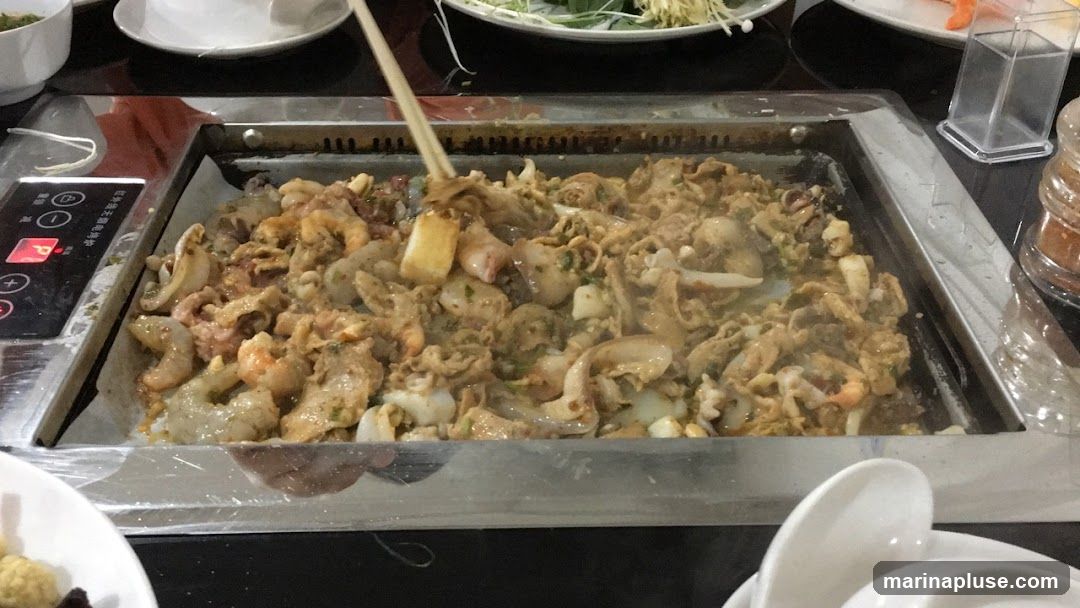 A close-up shot of a metal grill filled with stir-fried seafood and meat. Shrimp, squid, and other ingredients are visible, coated in a savory sauce and being stirred with chopsticks. The grill is part of a tabletop cooking setup, with a control panel on the left side. In the foreground, white spoons are stacked in a bowl. Other dishes and condiments are partially visible in the background.