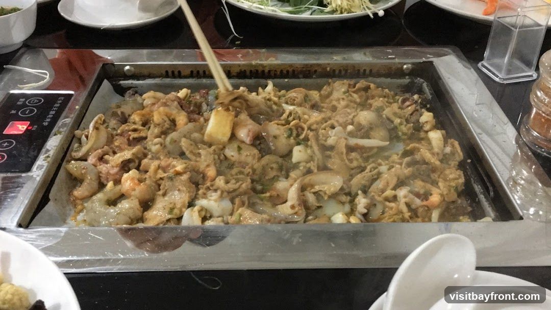 A close-up shot of a metal grill filled with stir-fried seafood and meat. Shrimp, squid, and other ingredients are visible, coated in a savory sauce and being stirred with chopsticks. The grill is part of a tabletop cooking setup, with a control panel on the left side. In the foreground, white spoons are stacked in a bowl. Other dishes and condiments are partially visible in the background.