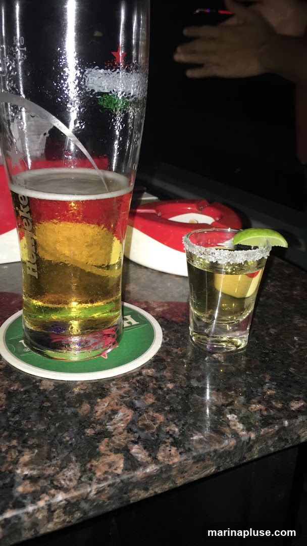 A pint of beer and a shot of tequila with a lime wedge sit on a granite countertop. The beer glass has a Heineken logo visible. In the background, a red and white ashtray is visible, and hands are clapping.