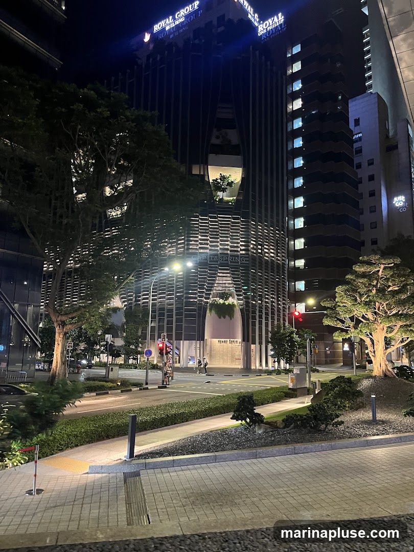 A nighttime view of a modern skyscraper with distinctive architectural features, including a curved facade and illuminated windows. The building is surrounded by trees and landscaping, with a street and sidewalk in the foreground. The "ROYAL GROUP" signage is visible at the top of the building.