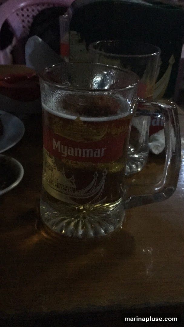 A glass mug filled with beer sits on a wooden table. The mug has a red label with the word "Myanmar" in white lettering, along with an illustration of a boat. Another glass is visible in the background.