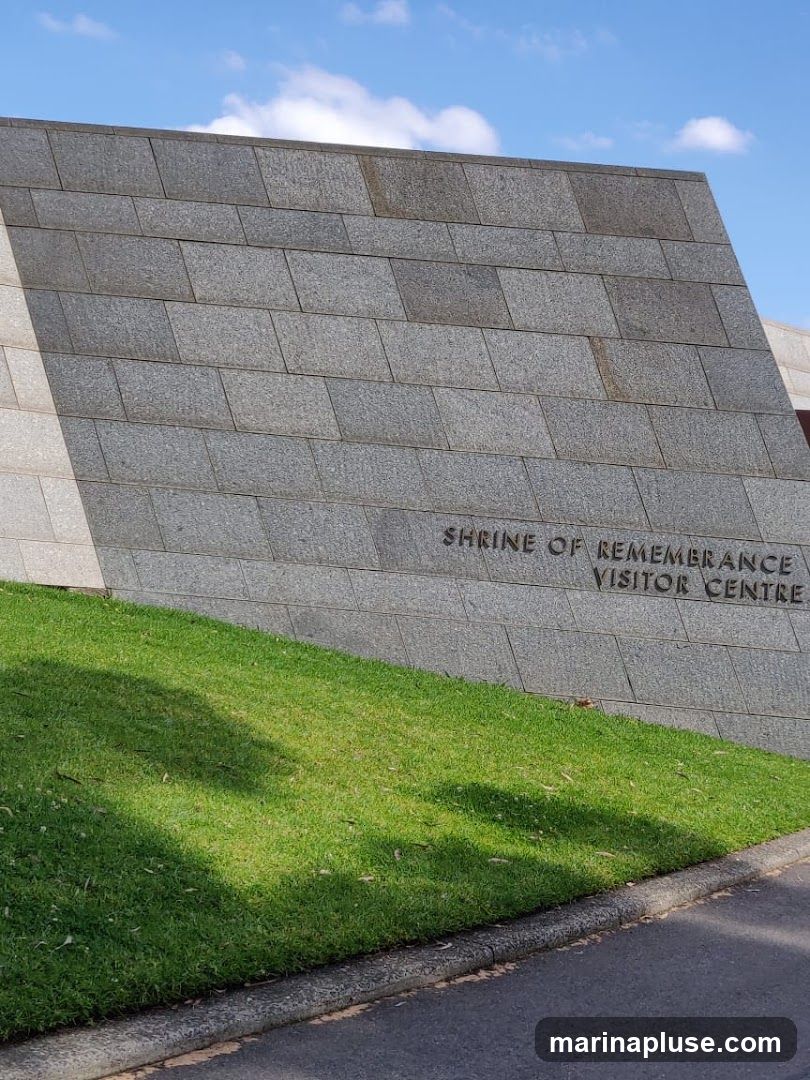 A close-up, low-angle shot of the exterior of the Shrine of Remembrance Visitor Centre. The building is clad in large, grey granite tiles. Text carved into the tiles reads "SHRINE OF REMEMBRANCE VISITOR CENTRE". A grassy slope is in the foreground, with a paved area and curb to the right. The sky above is blue with scattered white clouds.
