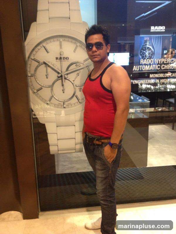 A man in a red tank top, blue jeans, and sunglasses stands in front of a large display of a Rado watch. Behind him, a Rado advertisement for the HyperChrome Automatic Chronograph is visible, along with display cases of watches.
