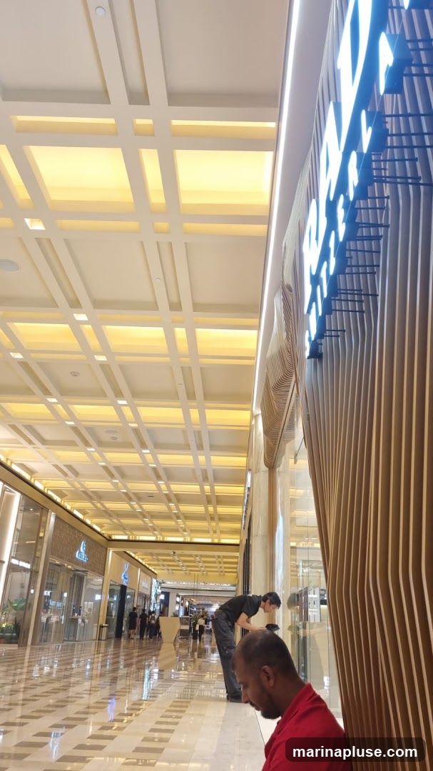 A wide shot of a brightly lit, modern shopping mall interior. The ceiling features a geometric design with recessed lighting. The floor is polished and reflective, showing the checkered pattern of the tiles. Several storefronts are visible in the background, with shoppers walking by. In the foreground, two men are present, one sitting and one leaning over a display. A large, illuminated sign with vertical wooden slats is on the right side of the frame.