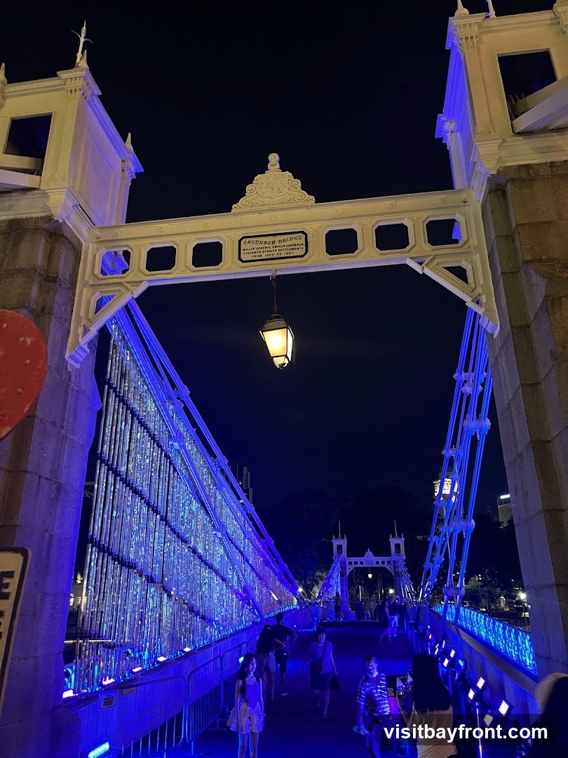 A view of Cavenagh Bridge at night, illuminated with blue lights. People are walking across the bridge. A lantern hangs from an archway above the bridge, which has a plaque that reads "CAVENAGH BRIDGE MAJOR GENERAL ORFEUR CAWLEY GOVERNOR OF THE STRAITS SETTLEMENTS FROM 1859 TO 1867".
