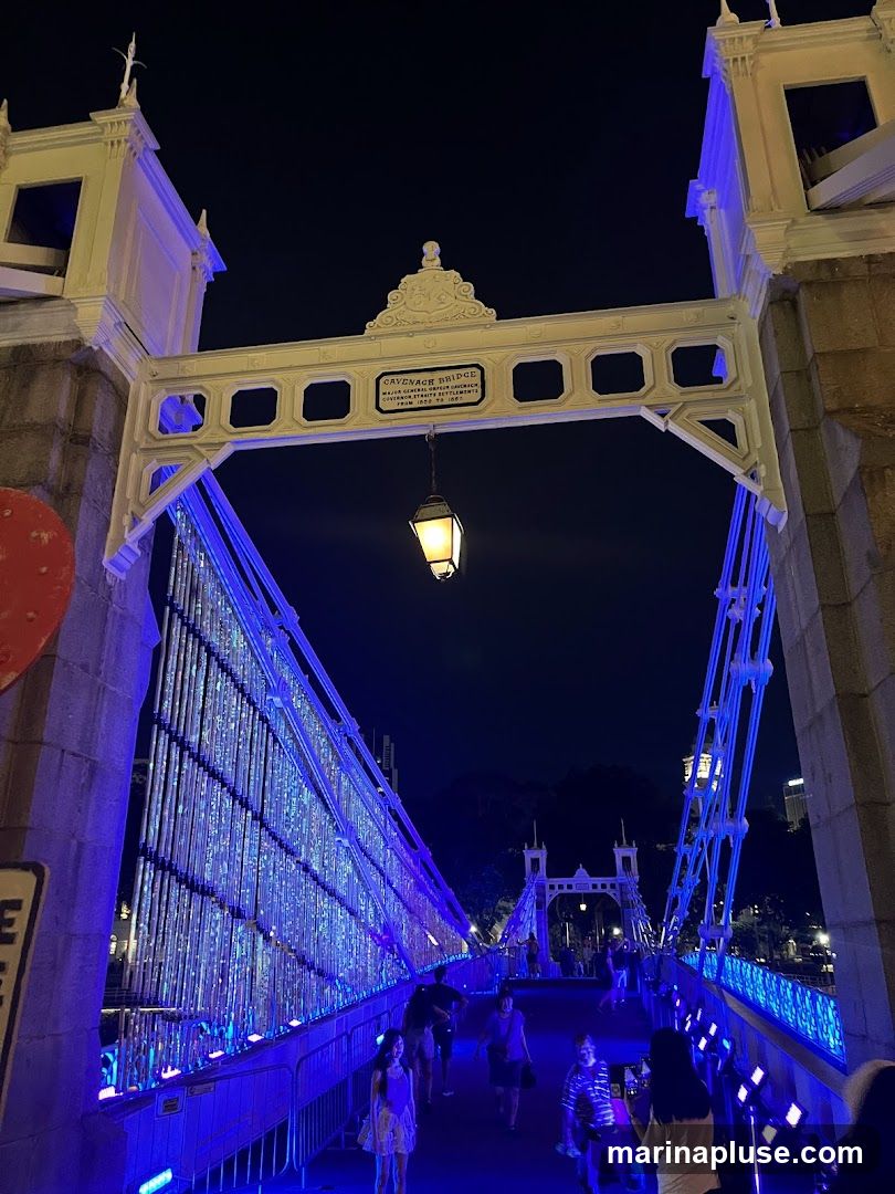 A view of Cavenagh Bridge at night, illuminated with blue lights. People are walking across the bridge. A lantern hangs from an archway above the bridge, which has a plaque that reads "CAVENAGH BRIDGE MAJOR GENERAL ORFEUR CAWLEY GOVERNOR OF THE STRAITS SETTLEMENTS FROM 1859 TO 1867".