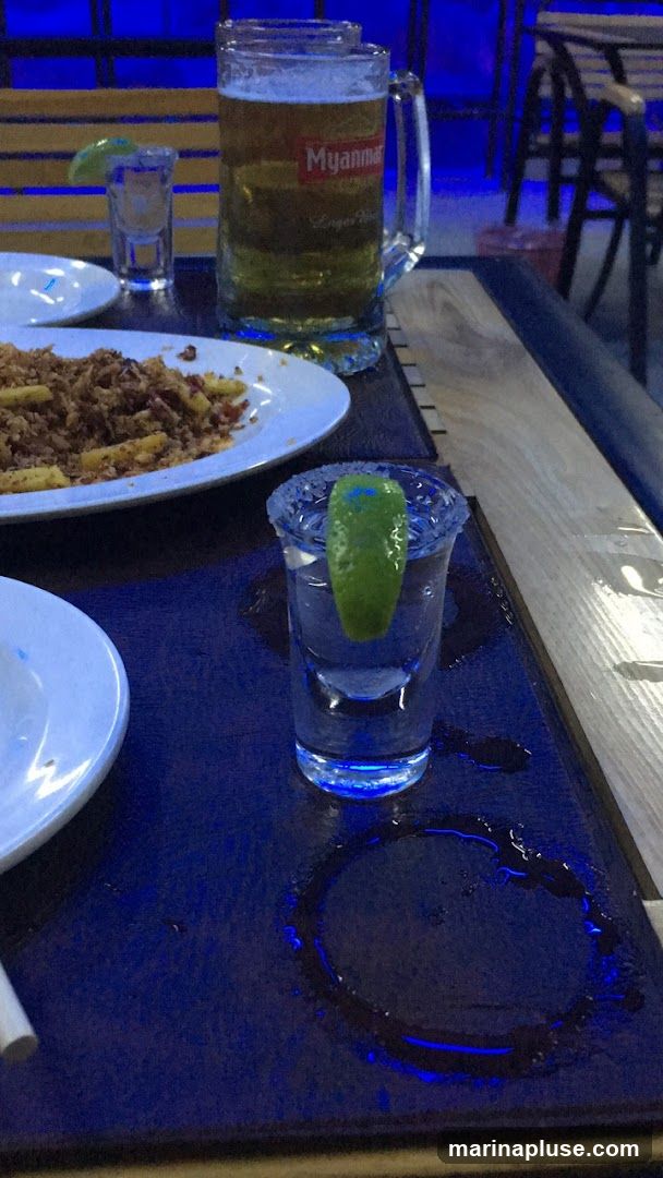 A close-up shot of a table with drinks and food. In the foreground, a shot glass with a lime wedge and a salt rim is prominently displayed. Behind it, a large mug of beer with a label that reads "Myanmar Lager Beer" is visible. A plate of food, possibly fried noodles or rice, is also on the table. The table surface is dark blue, and there are spills and condensation rings. In the background, blurred seating and blue lighting suggest an indoor or covered outdoor seating area.