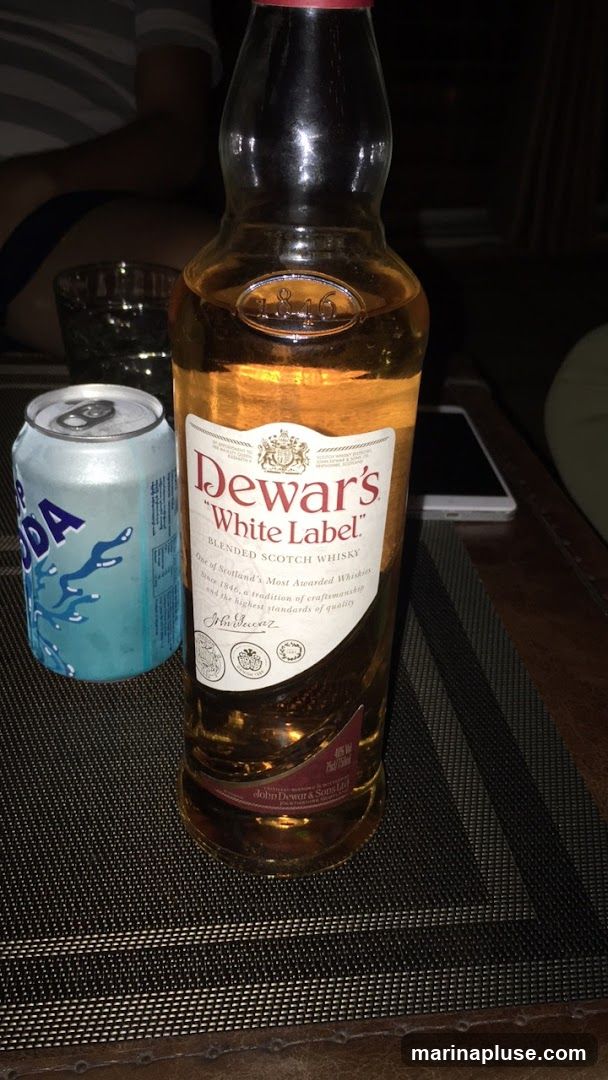 A bottle of Dewar's White Label Blended Scotch Whisky sits next to a can of soda on a table. The label on the bottle reads "Dewar's White Label Blended Scotch Whisky One of Scotland's Most Awarded Whiskies Since 1846, a tradition of craftsmanship and the highest standards of quality".