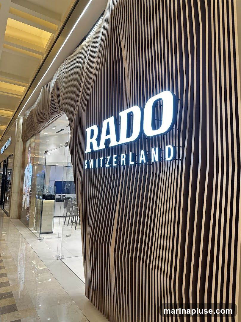 The exterior of a Rado watch store. The storefront features a modern design with vertical wooden slats that create a wave-like pattern. A large, illuminated RADO SWITZERLAND sign is prominently displayed. Through the glass entrance, display cases and seating are visible inside.
