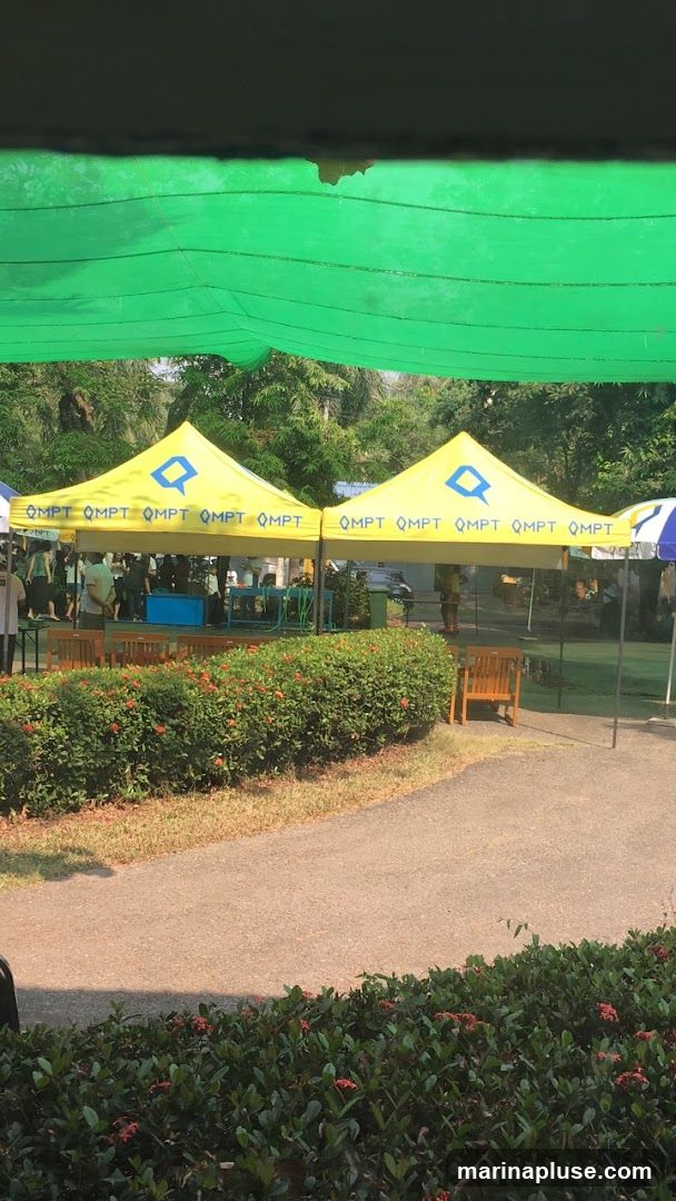 Two yellow tents are set up outdoors, with a green mesh covering overhead. The tents are branded with the text "QMPT" repeated multiple times and a blue logo resembling a stylized 'Q'. People are visible under the tents, and there are tables and chairs set up. A pathway and flowering bushes are in the foreground.