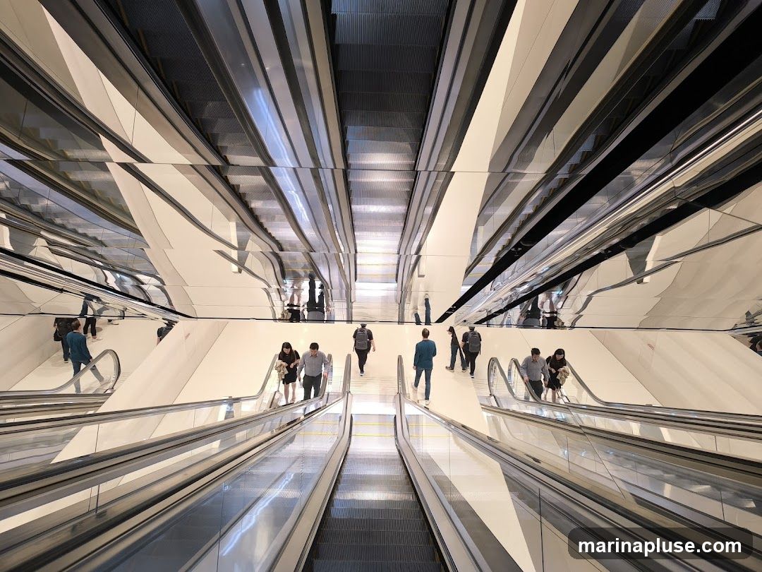 A wide-angle shot looking down a series of escalators in a modern building. The escalators are flanked by white walls and have metallic handrails. The ceiling is covered in mirrored panels, reflecting the escalators and the people using them, creating a disorienting and visually striking effect. Several people are seen ascending and descending the escalators.