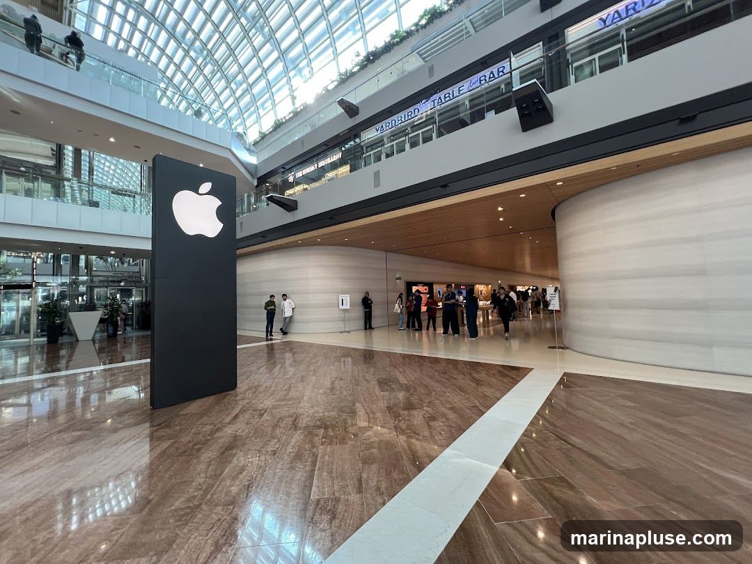 A large black pillar with a white Apple logo stands in the foreground of a modern indoor shopping mall. In the background, people are seen entering and browsing an Apple Store. Other shops, including one labeled "YARDBIRD TABLE AND BAR", are visible on upper levels. The mall features a glass ceiling and polished marble floors.