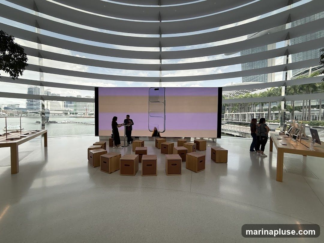 An interior view of a modern Apple Store with large windows overlooking a waterfront. A large screen displays a product image, and several wooden stools are arranged in a semi-circle in front of it. Display tables with Apple products are visible on either side of the room. People are browsing the products and interacting with staff.