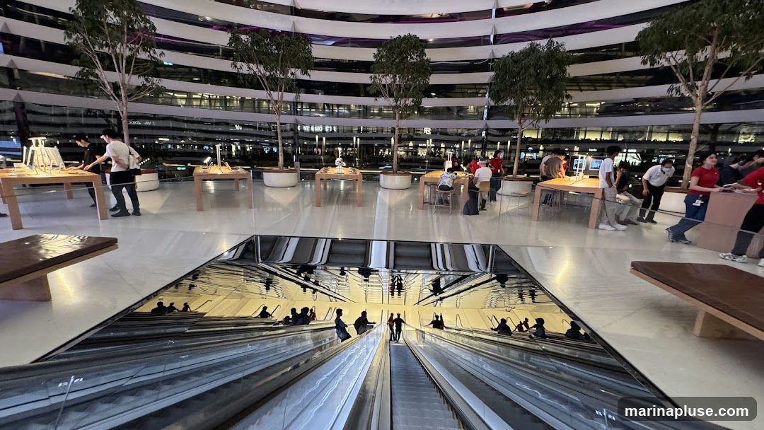 A spacious, modern interior with a reflective floor displaying an escalator scene. Several trees are planted in large white pots, adding greenery to the environment. People are seen browsing at tables displaying products, and staff members in red shirts are assisting customers. The background shows multiple levels of the building with reflective surfaces and lights.