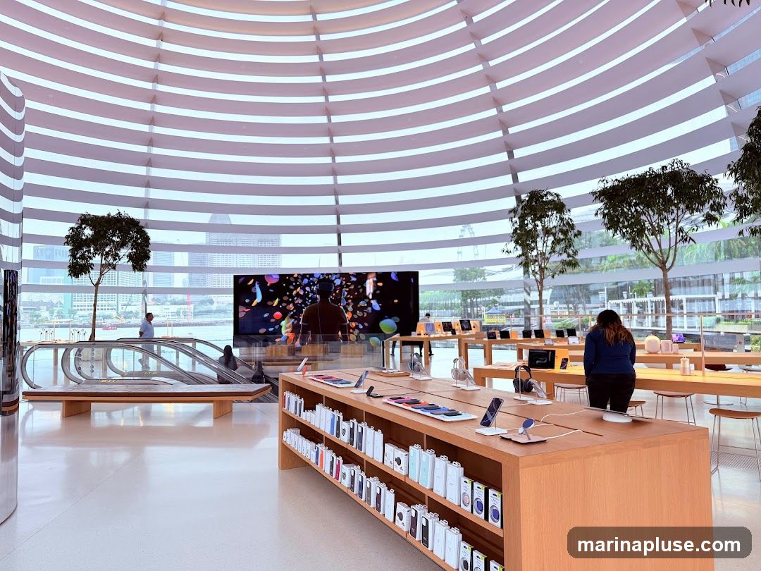 An interior view of a spacious, modern electronics store with a distinctive curved ceiling. Display tables showcase various electronic devices and accessories. An escalator is visible on the left, and large windows offer a view of the city skyline and trees outside. A large screen displays vibrant abstract graphics.