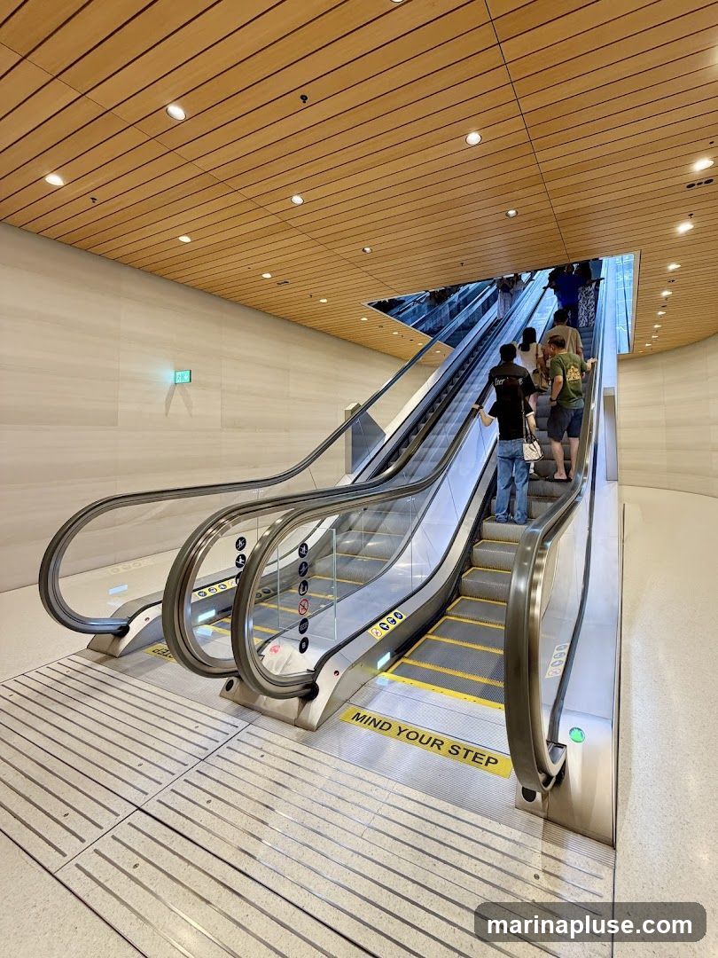 People are seen ascending on escalators in a contemporary building with a wooden slatted ceiling and marble-like walls. Yellow safety markings and "MIND YOUR STEP" signs are visible on the escalators. An illuminated green exit sign is mounted on the wall.