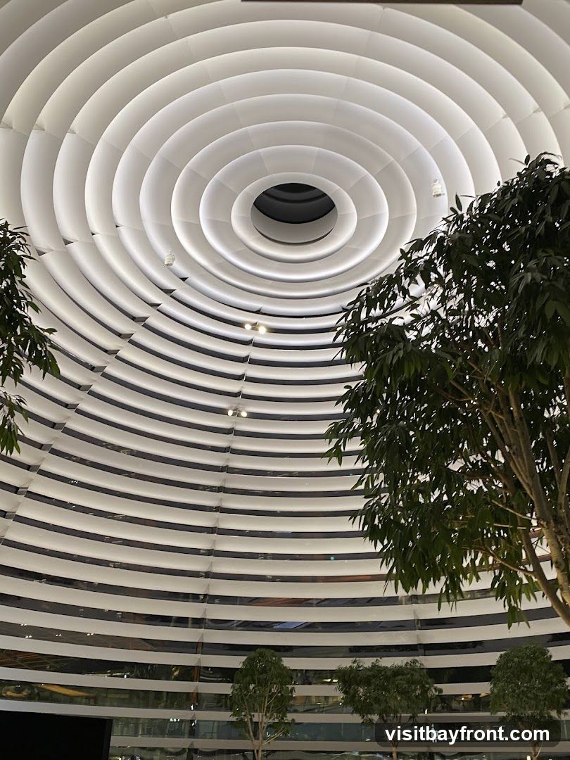 An interior view looking up at a striking modern ceiling composed of concentric white rings, illuminated from within. The rings create a sense of depth and a futuristic aesthetic. Several trees with lush green foliage are positioned within the space, adding a natural element to the contemporary design. The lower portion of the image shows reflections in glass, suggesting a large indoor area like a mall or atrium.