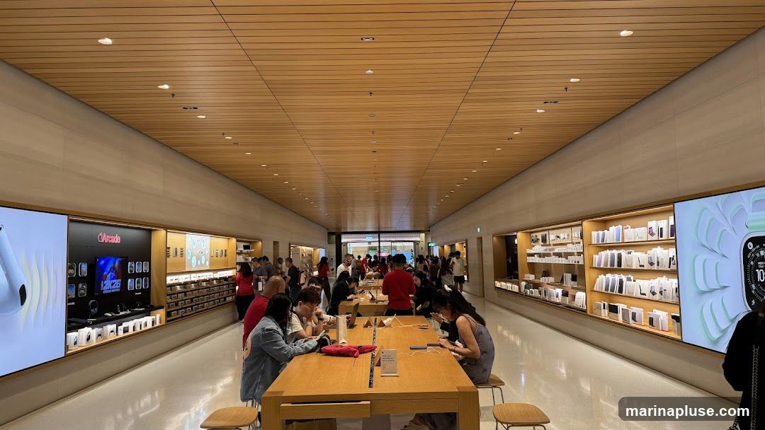A wide shot of the interior of an Apple Store, showing rows of wooden tables where customers are interacting with products. Shelves on the walls display various Apple products in their packaging. Large screens on the walls showcase product imagery. The ceiling is made of wood panels with recessed lighting. Several customers are seated at the tables, using iPhones and other devices.