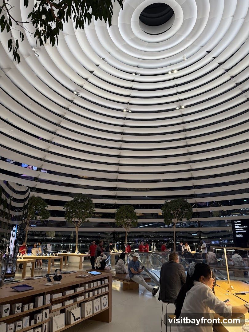 An interior view of a modern retail store with a distinctive domed ceiling featuring concentric white rings. The space is well-lit and features display tables with electronic products, including headphones and tablets. Several trees are placed throughout the store, adding a touch of nature. Customers are seen browsing and interacting with displays, while staff members in red shirts are also visible. The background shows a panoramic view through large windows, suggesting the store is located in a city with a waterfront.