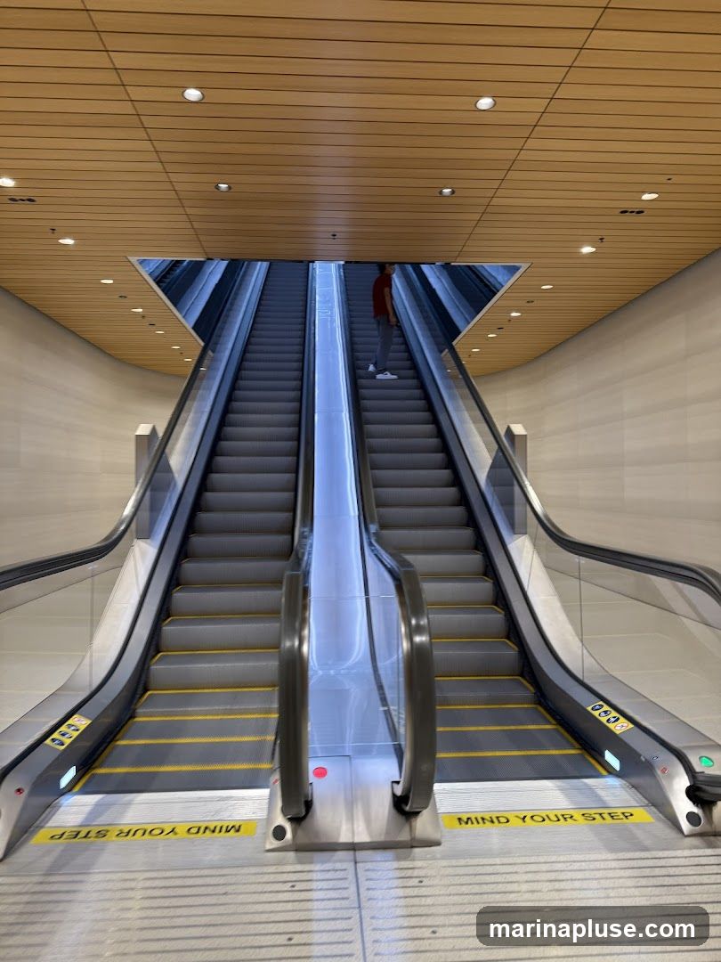 Two escalators ascend towards a triangular opening in a wood-paneled ceiling. A person in a red shirt stands on the right escalator. Yellow safety lines and "MIND YOUR STEP" warnings are visible on the escalator treads.