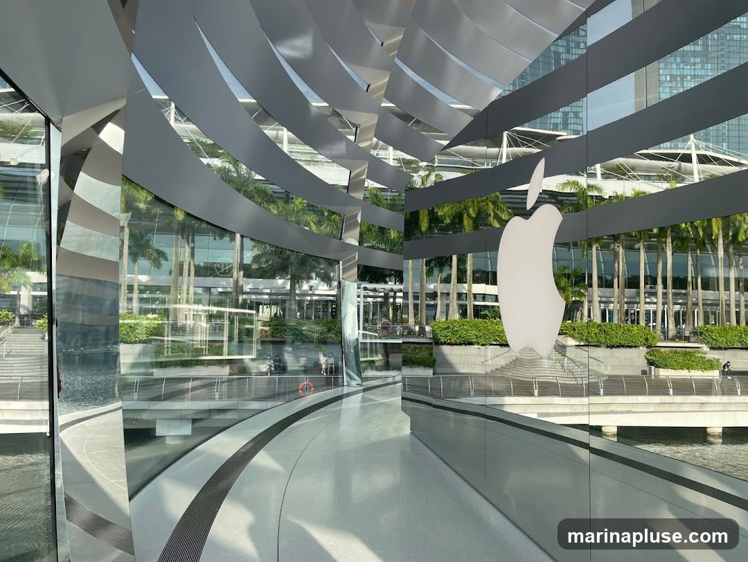 A large white Apple logo is displayed on a glass facade of a modern building. The building features a curved, reflective exterior with a metallic, slatted roof structure. Lush green palm trees and other vegetation are visible through the glass, along with reflections of the surrounding architecture and sky. In the background, other buildings and a body of water can be seen.