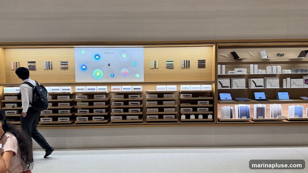 A wide shot of an Apple Store interior, showcasing various products and accessories. The left side features a display of Apple Watch bands arranged in rows on shelves, with a large screen above displaying Apple Watch interface elements. The right side of the store displays iPads, accessories like cases and styluses, and laptops. Several iPads are on display, showing their screens. The overall aesthetic is clean and modern, with light wood accents and bright lighting.