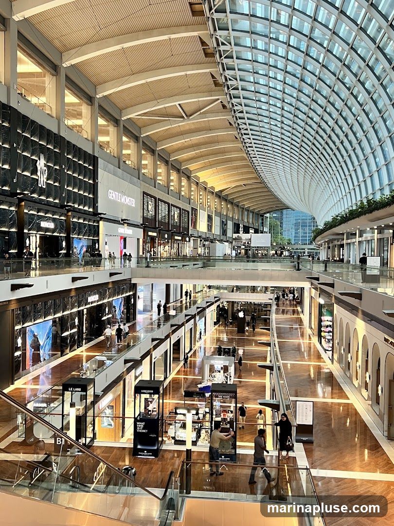 A wide-angle view of the interior of a large, modern shopping mall. The mall features a distinctive curved glass ceiling that allows natural light to flood the space. Multiple levels of shops are visible, with escalators and walkways connecting them. The shops have sleek, contemporary storefronts, some displaying illuminated signage for brands like Moncler and Gentle Monster. The flooring is polished wood, reflecting the lights and architecture. Several shoppers are seen browsing and walking through the mall.
