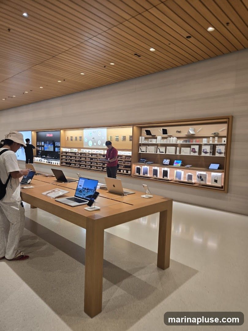 A well-lit electronics store interior with a long wooden table displaying laptops and phones. Behind the table, shelves are stocked with various electronic devices and accessories, including iPads and other Apple products. Several customers are browsing the merchandise. The ceiling is made of wood panels with recessed lighting.