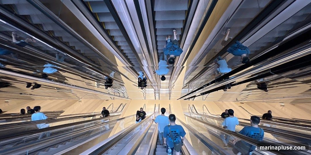 A wide-angle shot looking down a series of escalators, with mirrored ceilings creating a disorienting and expansive effect. Several people are visible riding the escalators in both directions. The escalators are flanked by metallic railings and the walls appear to be a light, textured material. The reflections in the ceiling show inverted images of the escalators and people, adding to the visual complexity.