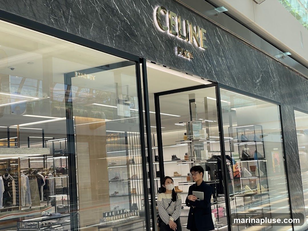 The exterior of a Celine store with the brand name "CELINE PARIS" displayed prominently above the entrance. The store features large glass windows showcasing merchandise, including clothing, handbags, and shoes. Two individuals, a woman wearing a mask and a man holding a tablet, are standing near the entrance, appearing to be staff or customers. The store's interior is visible through the windows, with shelves displaying various luxury goods.