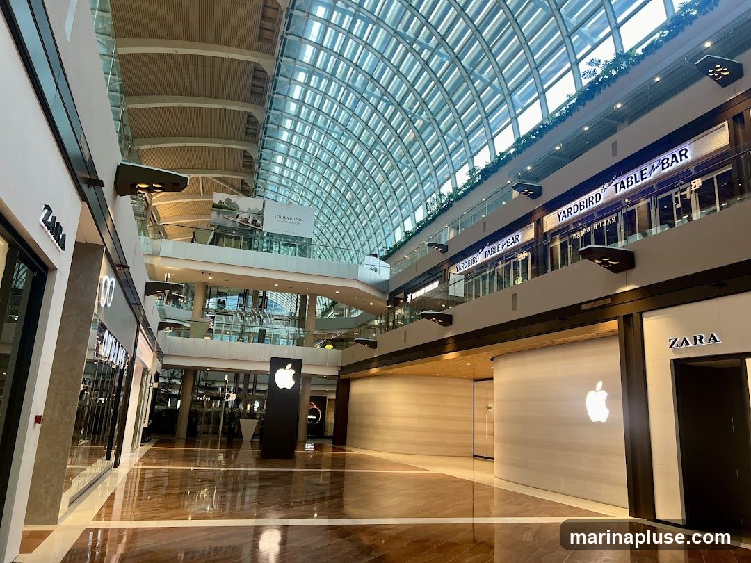 The image shows the interior of a large, modern shopping mall with a distinctive glass roof. Several retail stores are visible, including Zara and an Apple Store with its iconic logo illuminated on a curved wall. Above, on an upper level, signs for "YARDBIRD TABLE & BAR" and "LOUIS VUITTON ISLAND MAISON" are visible. The polished wooden floor reflects the light from the ceiling and the store displays. The overall impression is one of a spacious and upscale shopping environment.