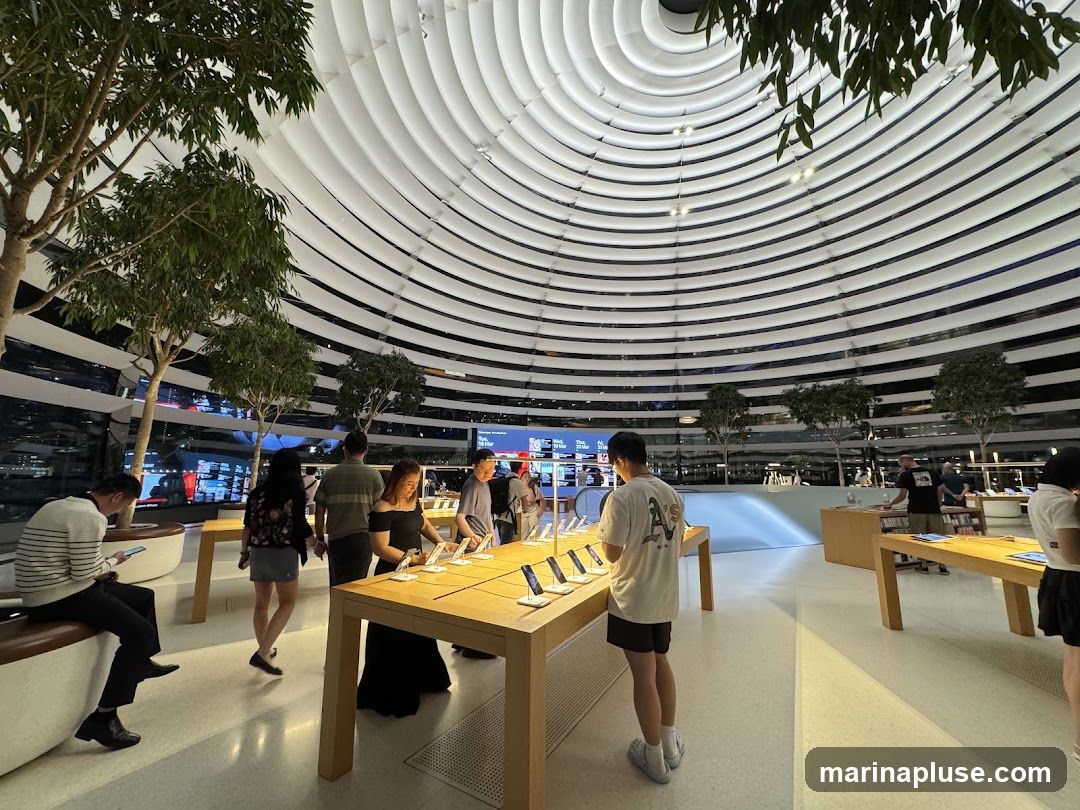 An interior view of a brightly lit, modern electronics store with a distinctive domed ceiling made of white ribbed panels. Several trees are placed strategically throughout the space, adding a touch of nature. Customers are browsing various electronic devices displayed on wooden tables. The atmosphere is clean, spacious, and contemporary.
