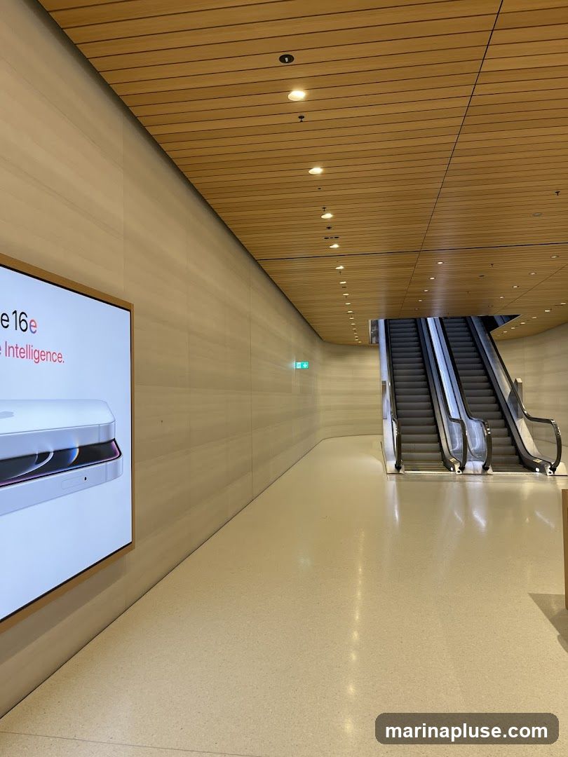 A wide shot of a modern interior space with a light-colored, speckled floor. To the left, a large digital display shows a close-up of a white electronic device with the text "e 16e" and "Intelligence." The walls are a light, textured material, and the ceiling is made of horizontal wooden slats with recessed lighting. In the background, a set of escalators ascends, with polished metal railings reflecting the light. A green exit sign is visible on the wall near the escalators.