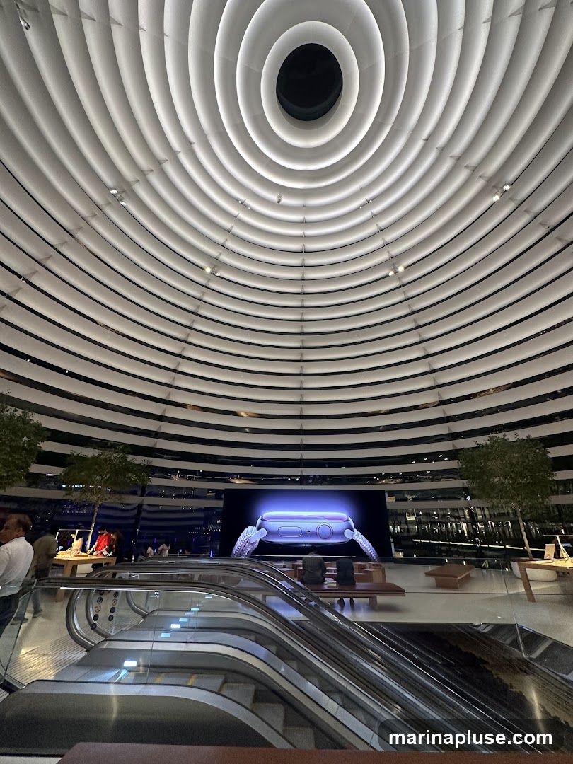 An interior view of a modern retail space with a distinctive circular, ribbed ceiling. In the center, a large screen displays a close-up of a purple smartwatch. Escalators are visible in the foreground, and several trees are placed strategically throughout the space. People are seen browsing and sitting on benches.