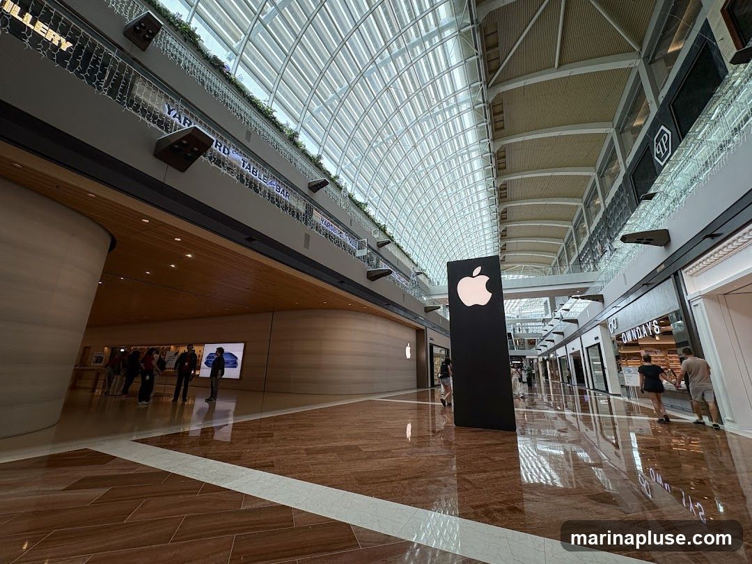 An interior view of a modern shopping mall with an Apple Store prominently featured. A large black sign with the Apple logo stands in the foreground. The mall has a high, arched glass ceiling and polished floors. Other storefronts are visible along the corridor, including one labeled "OWNDAYS". People are seen browsing inside the Apple Store and walking through the mall.