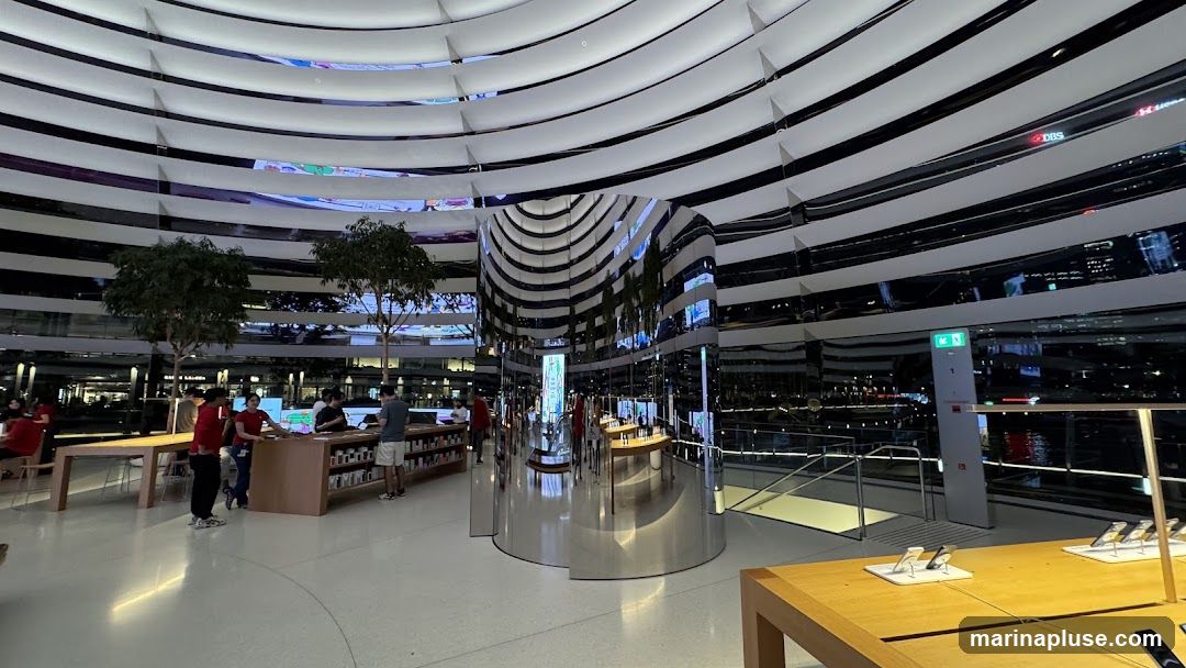 The interior of a spacious, modern retail store with a distinctive curved ceiling made of reflective white panels. Several trees are placed strategically throughout the space. In the foreground, wooden display tables showcase electronic devices. In the midground, staff members in red shirts interact with customers at counters. A large, cylindrical, mirrored pillar stands in the center, reflecting the surrounding environment. The background reveals a cityscape at night, visible through large windows.