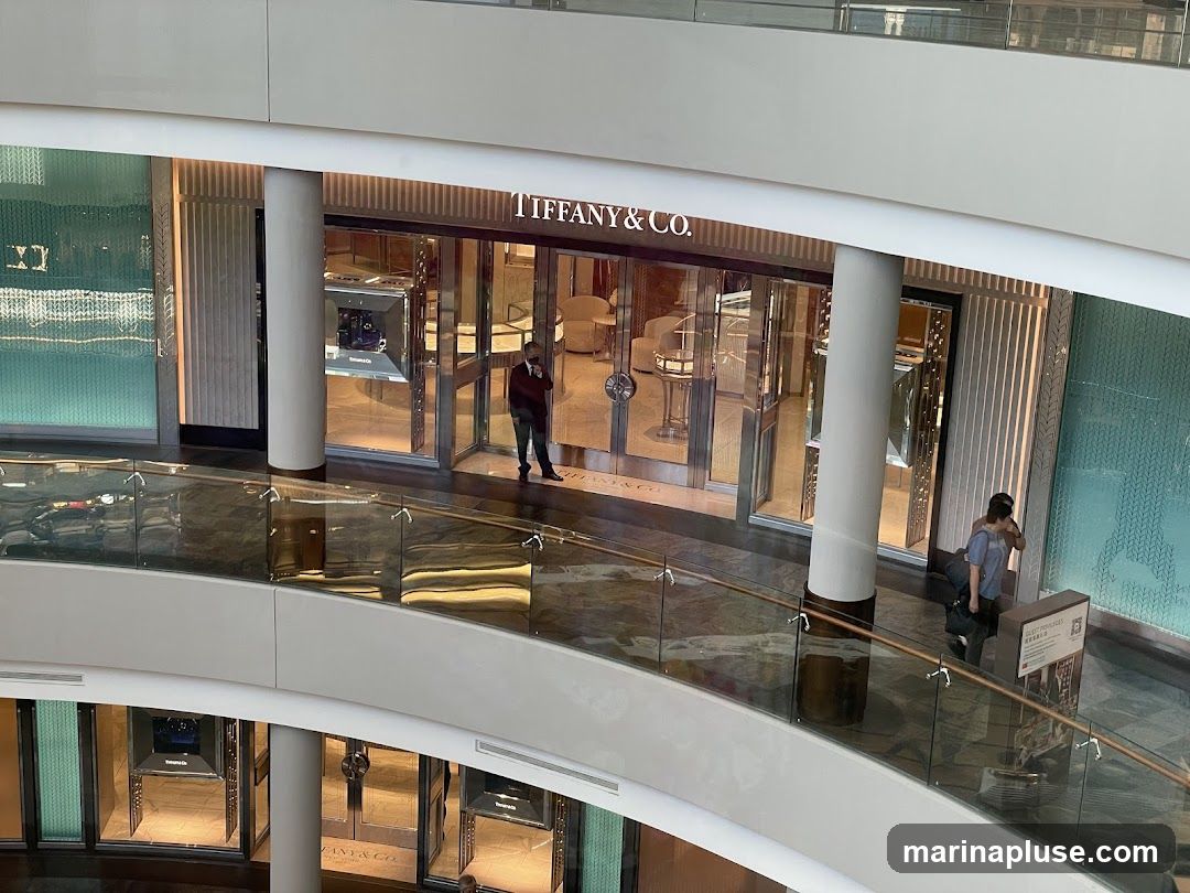 A high-angle view of the Tiffany & Co. storefront in a modern shopping mall. The entrance features large glass doors and windows, with the "TIFFANY & CO." logo prominently displayed above the entrance. Inside, display cases showcase jewelry and other luxury items. Two individuals are visible near the entrance, one standing and one seated. The surrounding mall architecture includes a curved walkway with glass railings and marble flooring.