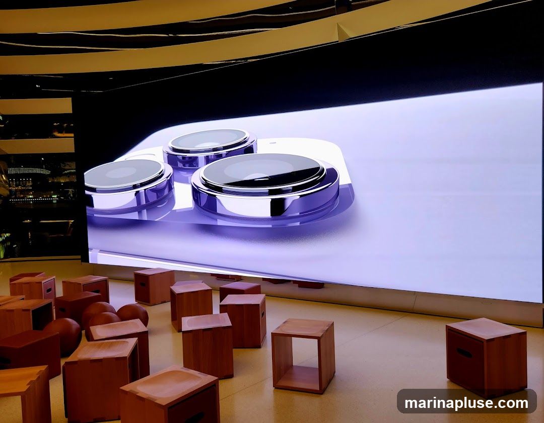 A large screen displays a close-up of a purple smartphone's camera lenses. In front of the screen, numerous wooden cube-shaped stools and some spherical objects are arranged on a light-colored floor. The ceiling features a modern, curved design with reflective elements.